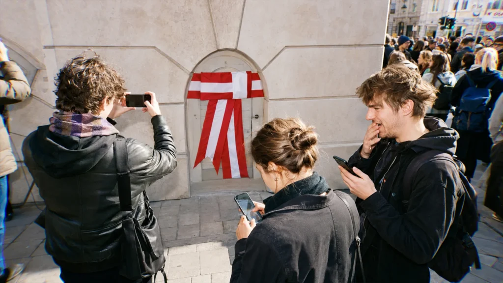 Drei junge Menschen dokumentieren während einer Wiener Demonstration eine absurde politische Inszenierung mit ihren Smartphones.