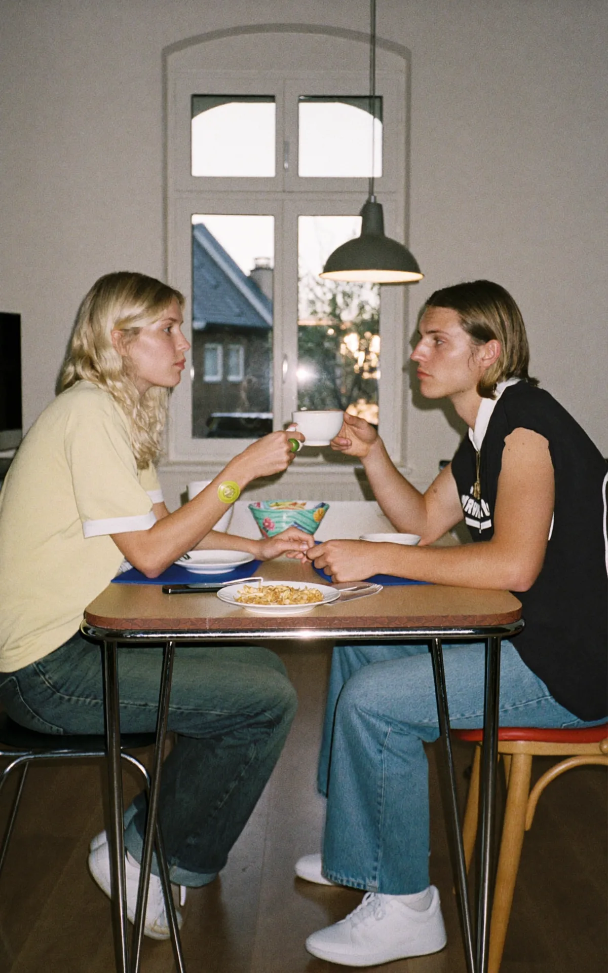 Zwei junge Partner sitzen nach einem Streit an einem Frühstückstisch, während eine Person der anderen vorsichtig eine Tasse zuschiebt.