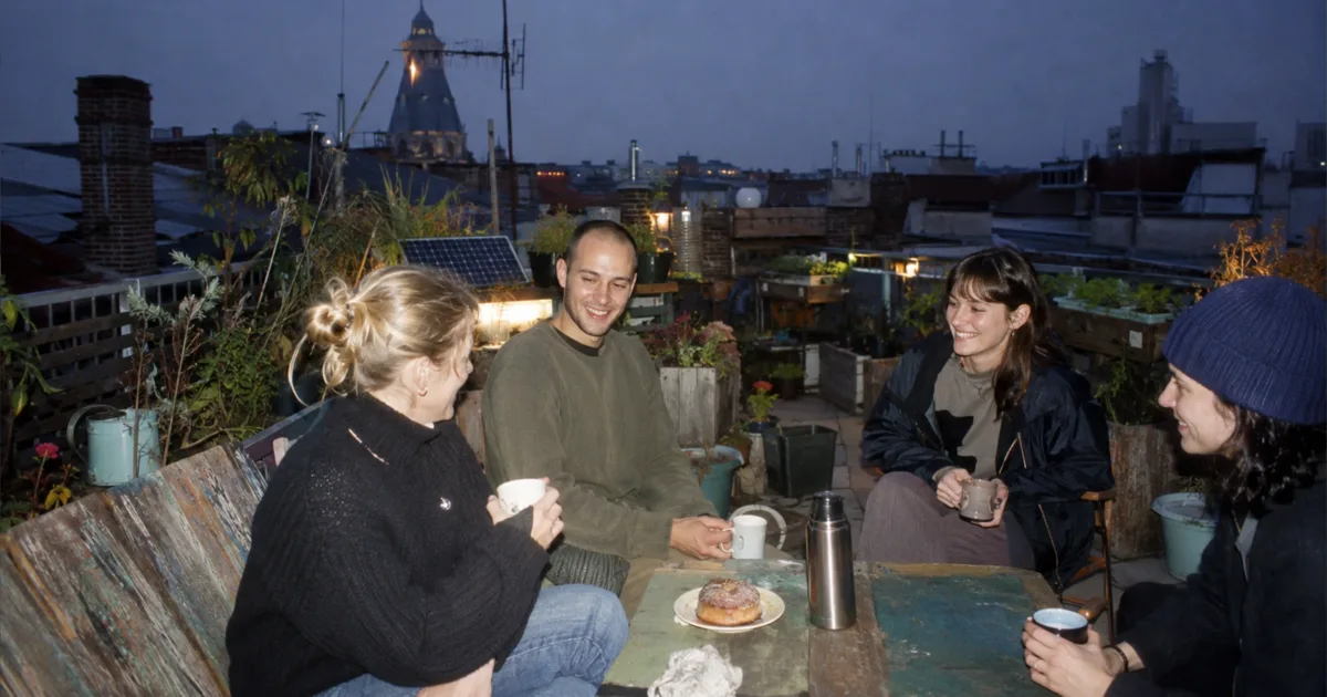 Junge Menschen teilen auf einem Wiener Gemeinschaftsdachgarten Kaffee und Gebäck zwischen reparierten Pflanzgefäßen.