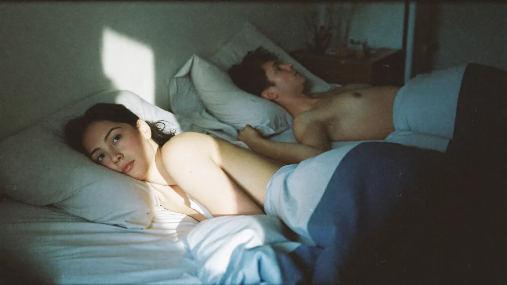 Eine Person liegt nach einem Streit wach im Bett, während der Partner auf der anderen Seite schläft.