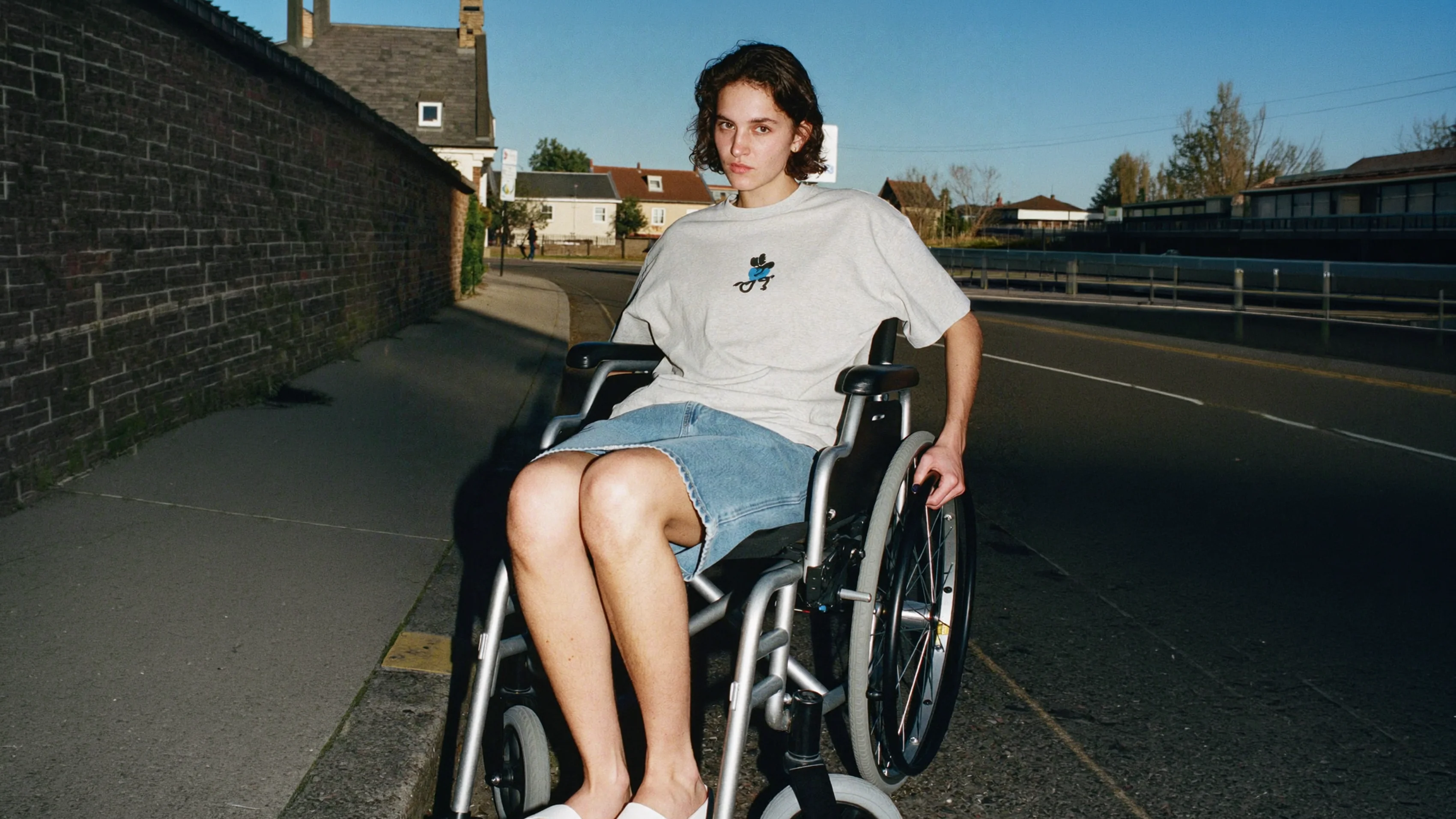 ine Frau mit kurzen, lockigen Haaren, die in einem manuellen Rollstuhl auf einem städtischen Gehweg sitzt. Sie trägt ein übergroßes graues T-Shirt mit einer blauen Cartoon-Grafik und einen Jeansrock. Sie ist neben einer alten Backsteinmauer positioniert, im Hintergrund eine Straße und Vorstadthäuser pod vedrim plavim nebom.