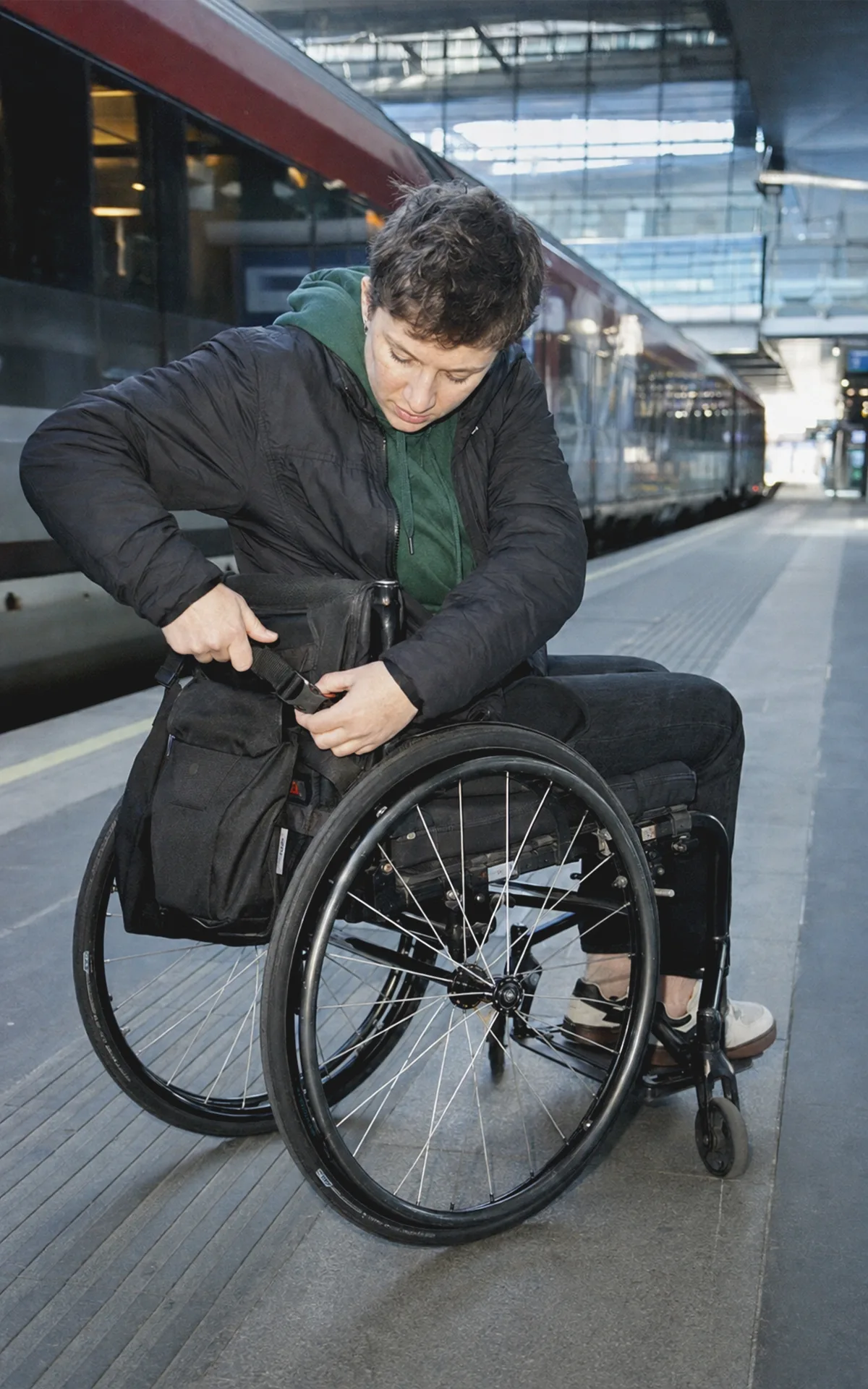Eine Person im Rollstuhl befestigt auf einem Wiener Bahnsteig selbstständig eine Reisetasche.