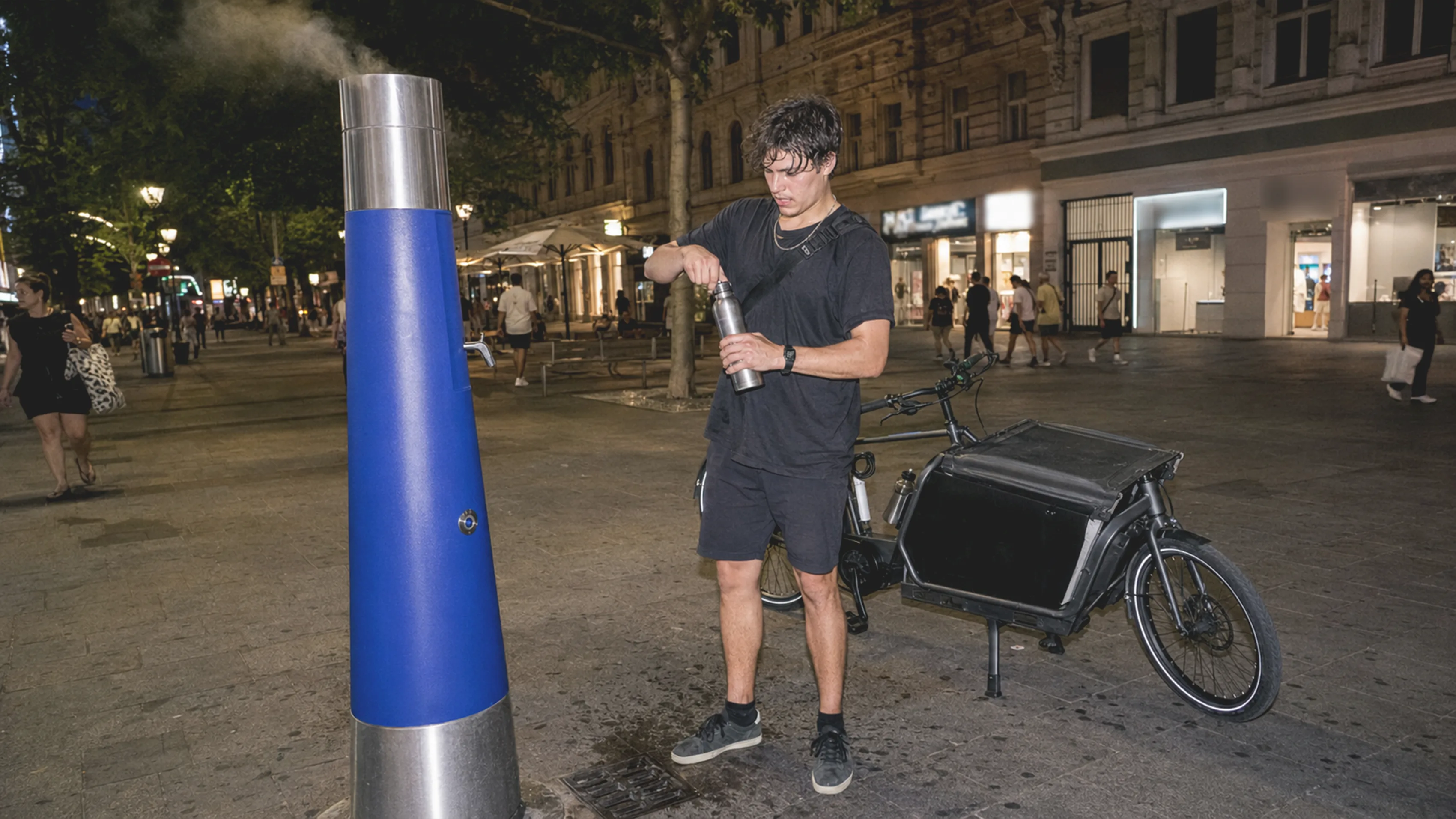 Alternative Text DE: Ein Fahrradkurier füllt an einer Brunnhilde auf der Wiener Mariahilfer Straße eine Wasserflasche auf.