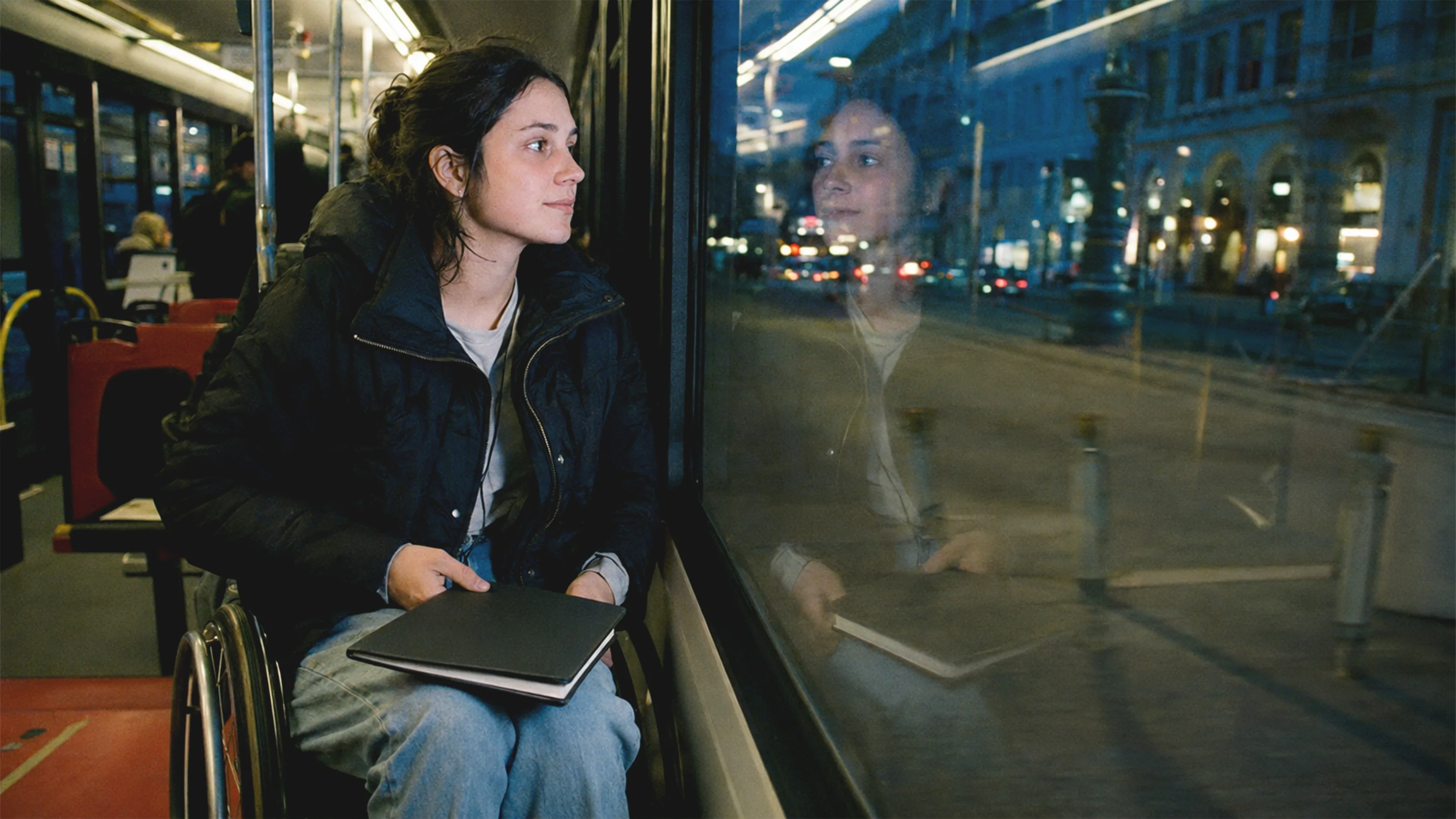 Eine junge Frau im Rollstuhl sitzt in einer Wiener Straßenbahn, während sich ihr Gesicht zwischen vorbeiziehenden Lichtern im Fenster spiegelt.