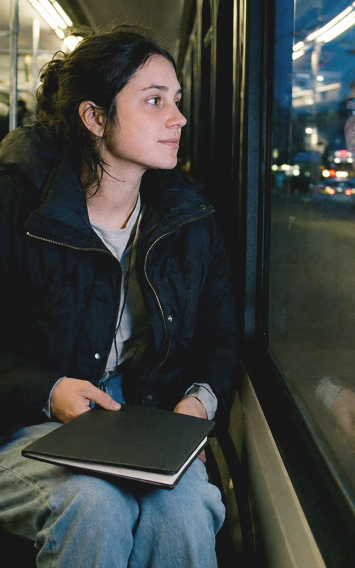 Eine junge Frau im Rollstuhl sitzt in einer Wiener Straßenbahn, während sich ihr Gesicht zwischen vorbeiziehenden Lichtern im Fenster spiegelt.